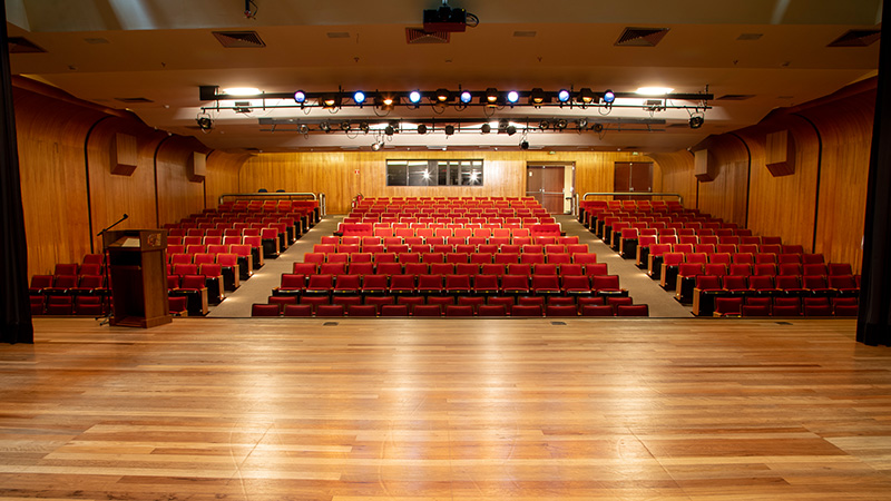 Foto tirada do palco, mostrando ao fundo as portas de acesso, janelas da sala de controle e as cadeiras vermelhas do Teatro CIEE