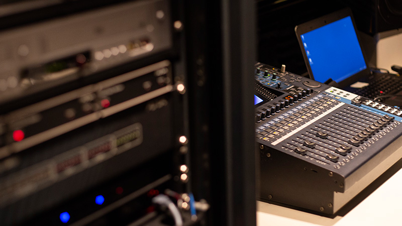 Foto de equipamentos na sala de controle do Teatro CIEE, mostrando ao fundo uma mesa com diversos botões e um notebook