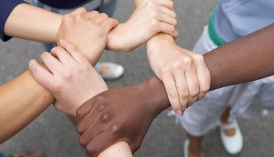 Destaque para cinco mãos de pessoas de diferentes etnias unidas vistas de cima.