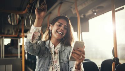 Moça jovem em pé dentro de um ônibus de dia segura celular em uma das mãos e sorri
