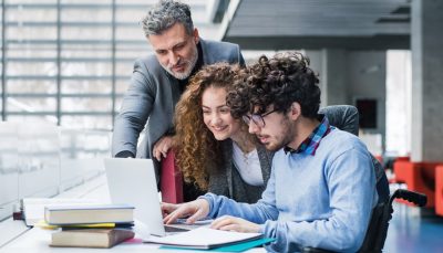 Estudante cadeirante em frente ao notebook e ao seu lado estão um homem e uma jovem