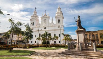 Imagem de praça com igreja branca colonial ao fundo em dia ensolarado