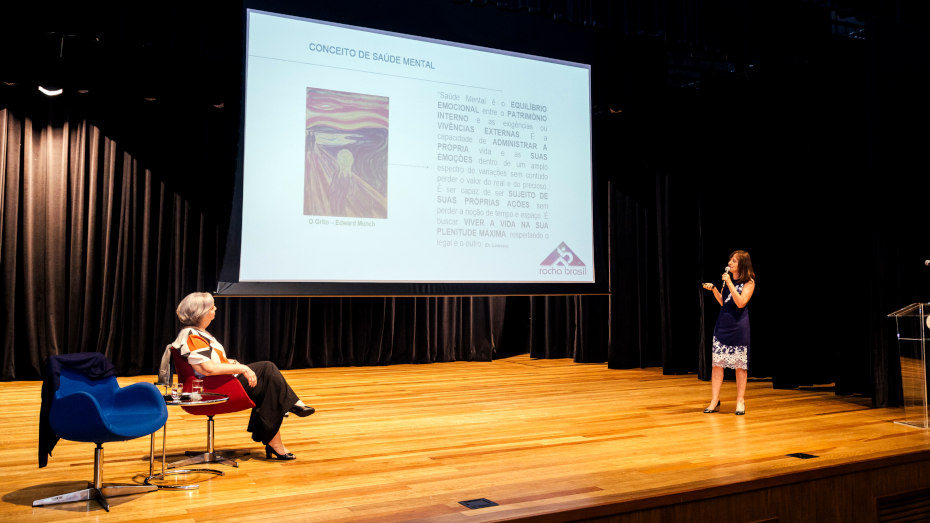 Duas mulheres em palco, uma sentada em cadeira enquanto a outra faz apresentação de slides em telão
