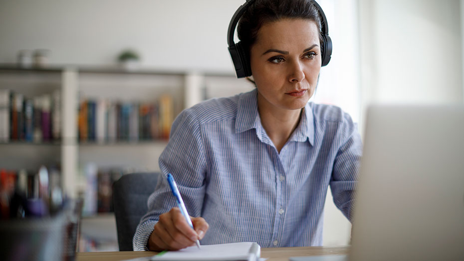 Mulher trabalhando com fone de ouvido, olhando para o computador e fazendo anotações