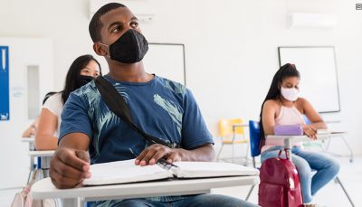 Em destaque um rapaz sentado em sala de aula usando máscara facial e duas meninas logo atrás também com máscara