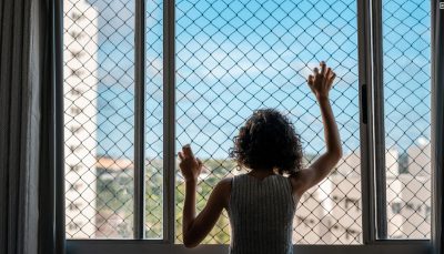 Menina, de cabelo encaracolado, está com as duas mãos apoiadas em uma tela da janela de aparamento