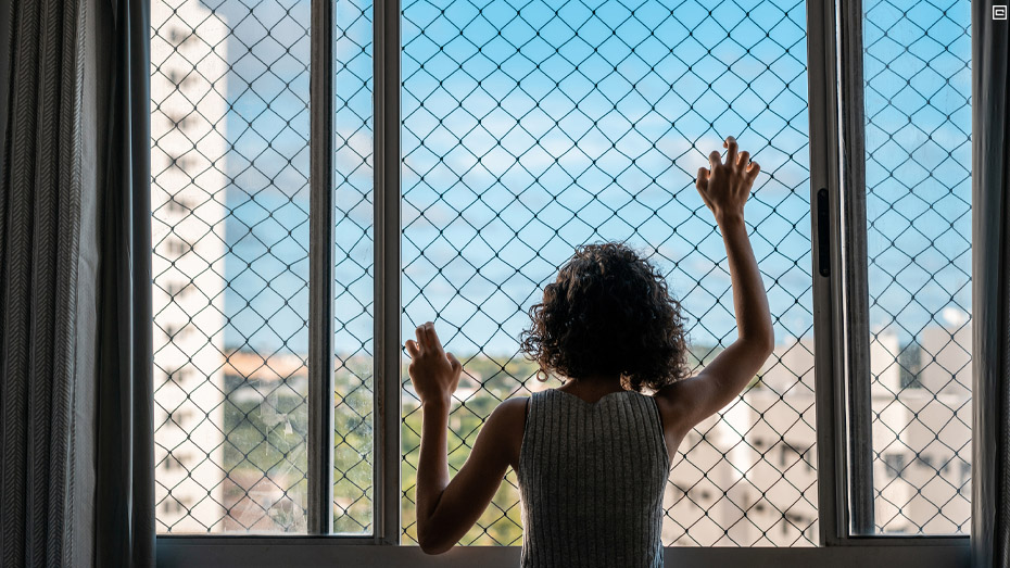 Menina, de cabelo encaracolado, está com as duas mãos apoiadas em uma tela da janela de aparamento