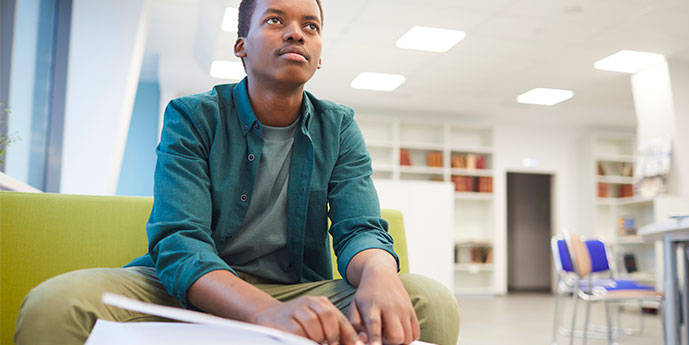 #PraCegoVer #ParaTodosVerem #DescriçãoDaImagem: Foto de um homem jovem negro em um ambiente escolar.  Ele olha para frente, usa uma camisa azul e uma calça bege. Está sentado em um sofá e na sua frente tem um livro sobre régua e faz leitura braille. Fim da descrição.