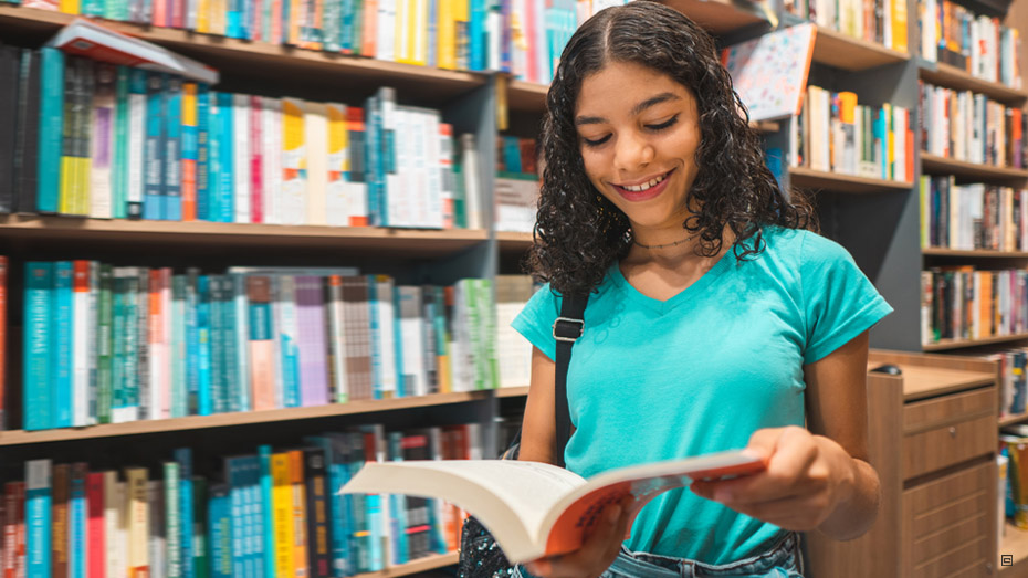 Uma jovem morena de cabelos encaracolados e com mochila preta nas costas sorri com um livro aberto em suas mãos. Ao fundo várias estantes de livro.