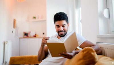 Homem preto, de cabelo encaracolado e barca, está sentado no sofá de sua casa com um livro aberto nas mãos
