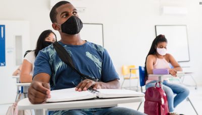 Estudante usando máscara sentado em uma poltrona universitária, ao fundo duas estudantes também usando máscara