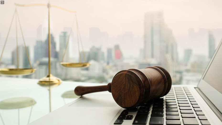 Martelo da justiça sobre o teclado de um notebook, com balança dourada e prédios ao fundo