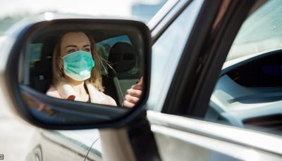 Mulher loira, com camisa rosa, está dirigindo um carro e é possível ver sua imagem refletida no retrovisor