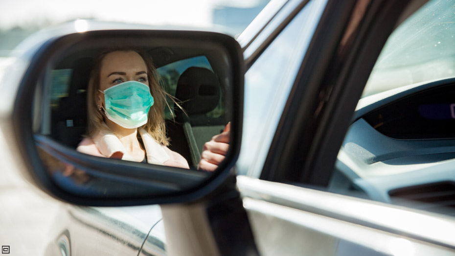 Mulher loira, com camisa rosa, está dirigindo um carro e é possível ver sua imagem refletida no retrovisor