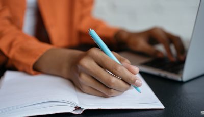 Imagem destaca mão de uma mulher negra escrevendo em um caderno sobre uma mesa, ao fundo é possível ver que ela está com a outra mão em um notebook