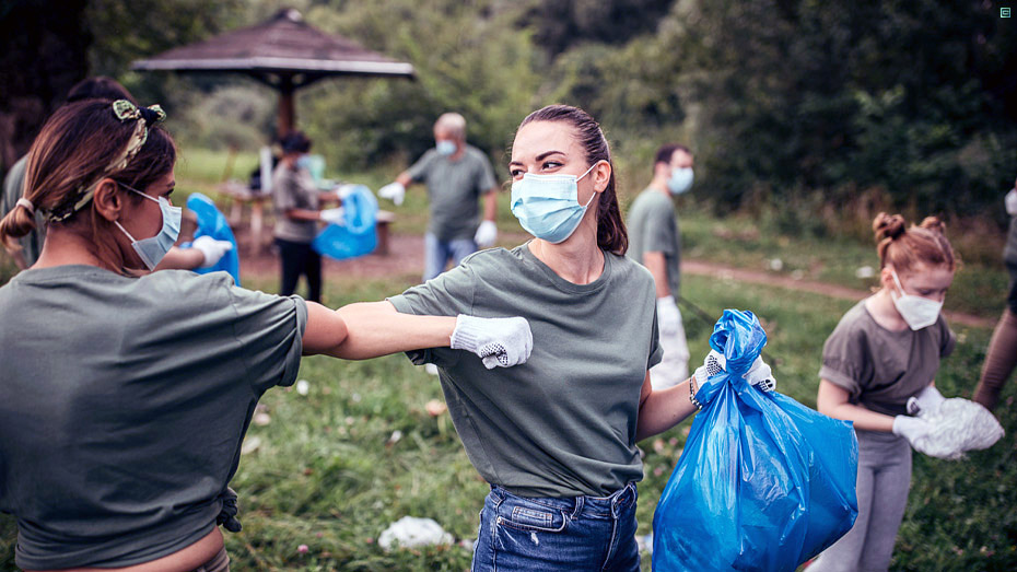 Pessoas recolhendo lixo de um local gramado, destaque para duas mulheres se cumprimentando tocando os ombros