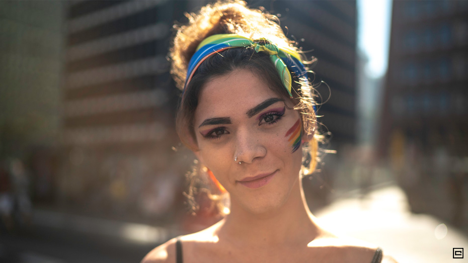 Pessoa trans morena sorrindo com tiara colorida e bandeira do orgulho LGBTQIA+ pintada no rosto,