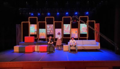 Palco da Expo CIEE Virtual com pufes coloridos e sofá branco. No centro do palco dois homens brancos e uma mulher negra