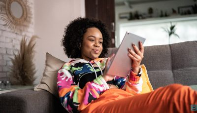 Jovem preta com os cabelos encaracolados pretos está sentada no sofá olhando um tablet que está em suas mãos