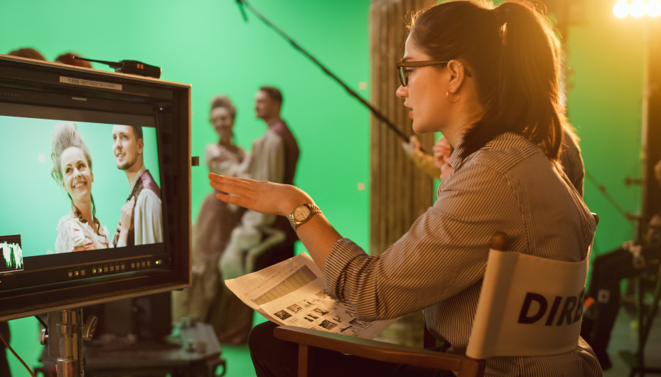 Mulher sentada na cadeira de diretora comanda uma cena de gravação enquanto vê atores na tela da câmera